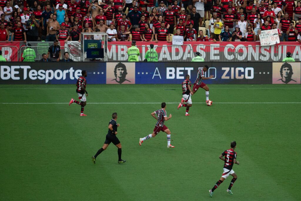 Imagem de um campo de futebol com jogadores correndo e placa da BETesporte ao fundo.